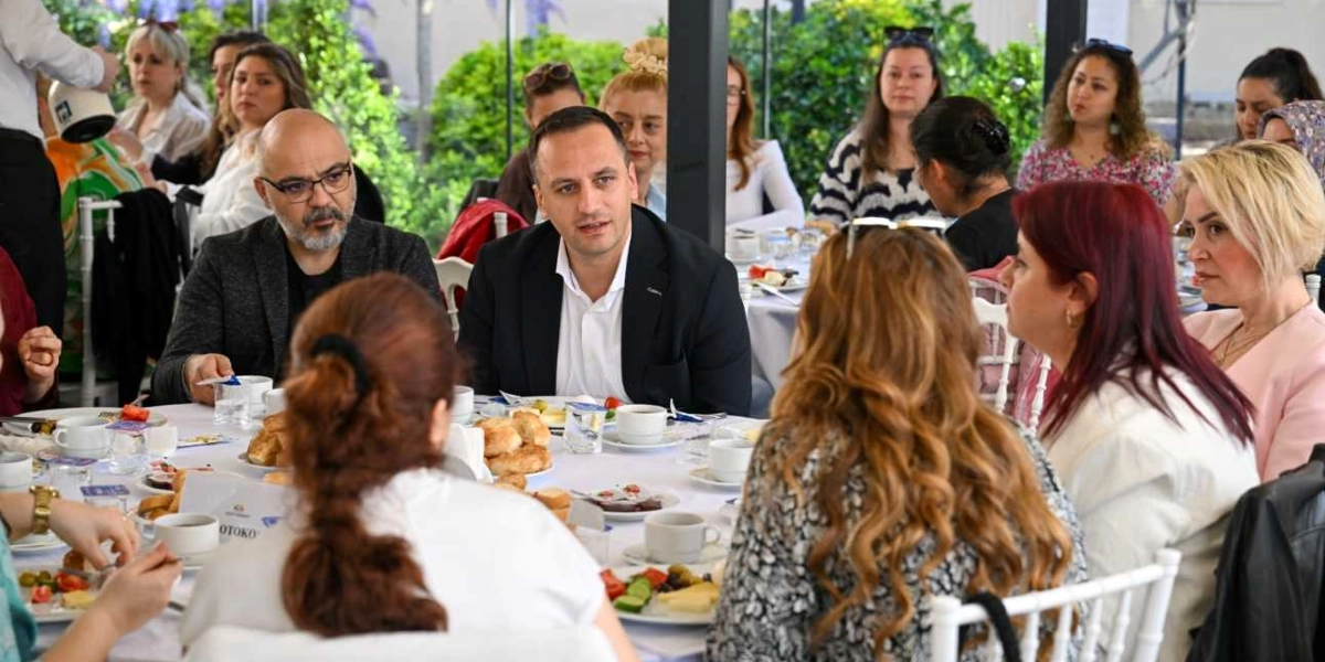 Açık havada, beyaz örtülü yuvarlak masaların etrafında toplanmış bir grup insan kahvaltı yaparken sohbet ediyor. Masanın merkezinde, koyu renk ceketli ve beyaz gömlekli bir erkek konuşma yaparken, çevresindeki katılımcılar onu dinliyor. Masalarda kahvaltılıklar, çay bardakları ve tabaklar bulunuyor. Çoğunluğu kadınlardan oluşan katılımcılar, samimi bir ortamda bir araya gelmiş durumda.