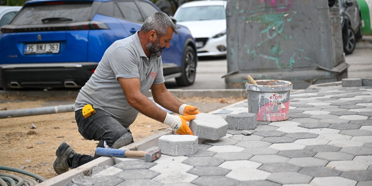 Gri polo yaka tişörtlü, sakallı bir işçi, Zafer Caddesi'nde altıgen kilit parke taşlarını döşeyerek kaldırım düzenlemesi yapıyor. İşçi, dizlerinin üzerinde çömelmiş durumda, ellerinde turuncu iş eldivenleri var ve bir taşı yerine yerleştiriyor. Yanında bir kova, çekiç ve mezura bulunuyor; arka planda ise park halindeki araçlar ve cadde ortamı görülüyor.