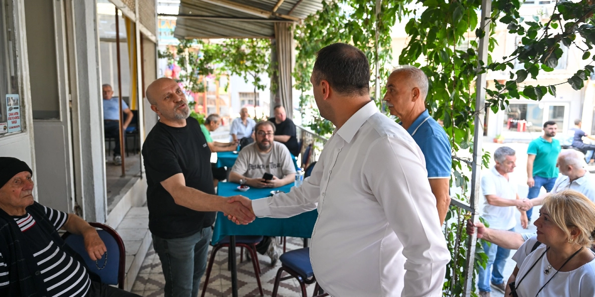 Beyaz gömlekli bir erkek, bir çay bahçesinde oturan siyah tişörtlü bir adamla tokalaşıyor. Çevredeki masalarda oturan ve ayakta duran diğer vatandaşlar bu anı izliyor. Ortam, sarmaşıklarla çevrili, yarı açık bir kafe veya çay ocağı görünümünde.