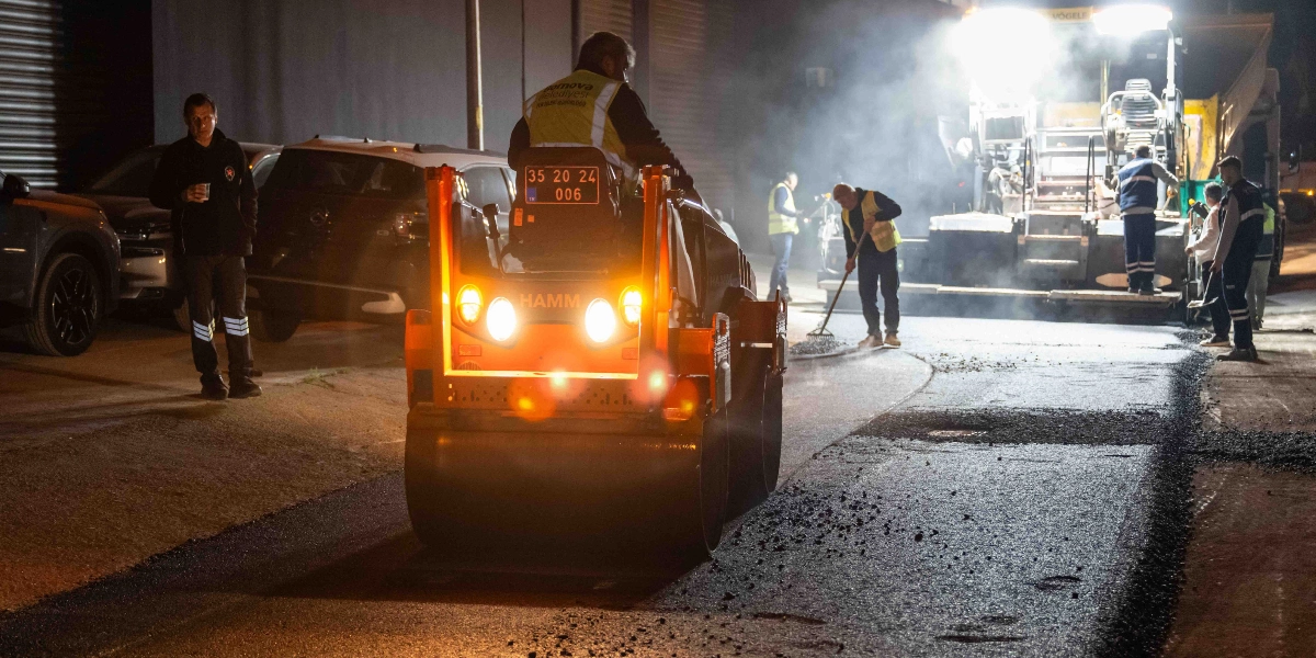 Gece vakti süren yol asfaltlama çalışmasında, üzerinde Bornova Belediyesi yazılı reflektörlü yelek giyen bir operatörün kullandığı Hamm marka silindir öne çıkıyor. Çalışma alanında işçiler tırmıklarla asfaltı düzeltirken, arka planda güçlü aydınlatmalarla donatılmış bir asfalt serici makine ve çevrede gözlem yapan kişiler bulunuyor.