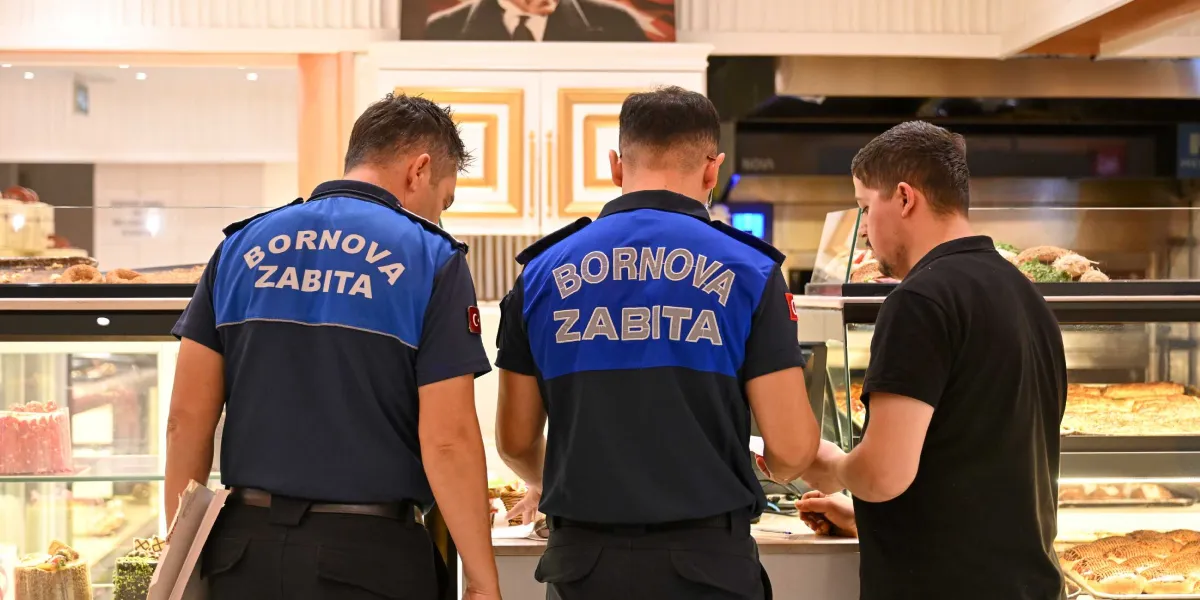 Sırtlarında Bornova Zabıta yazılı mavi-lacivert üniformalı iki zabıta memuru, bir fırın veya pastanede çalışan siyah tişörtlü bir kişiyle tezgah başında denetim yapıyor.