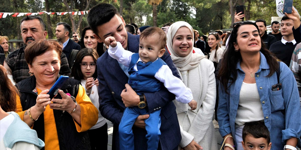 Açık havada düzenlenen bir etkinlikte, lacivert takım elbiseli genç bir adamın kucağında mavi yelekli ve papyonlu küçük bir erkek çocuk bulunuyor. Çocuğun yanında beyaz kıyafetli ve başörtülü bir kadın ile mavi gömlekli bir kadın gülümseyerek duruyor. Ön planda sarı bluzlu ve siyah yelekli bir kadın elindeki telefonla fotoğraf çekiyor, çevrede ise etkinliği izleyen kalabalık bir grup yer alıyor.