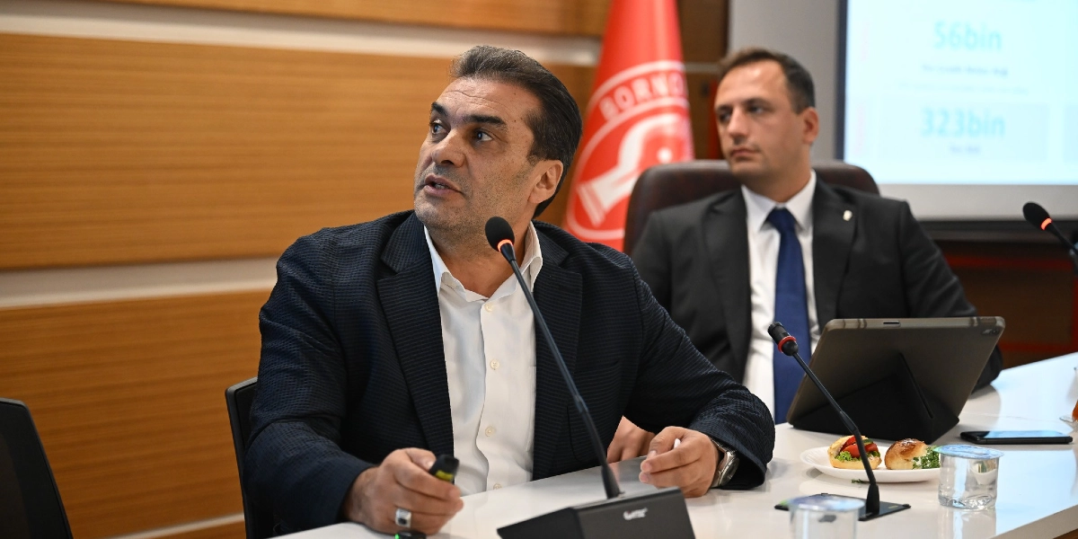Toplantı masasında oturan, lacivert ceketli ve beyaz gömlekli bir erkek konuşma yaparken, hemen arkasında takım elbiseli ve mavi kravatlı başka bir erkek onu dinliyor. Arka planda Bornova Belediyesi'nin logosunun yer aldığı kırmızı bir bayrak ve üzerinde verilerin bulunduğu bir sunum ekranı görünüyor. Masanın üzerinde mikrofonlar, bir tablet ve küçük atıştırmalıklar bulunuyor.
