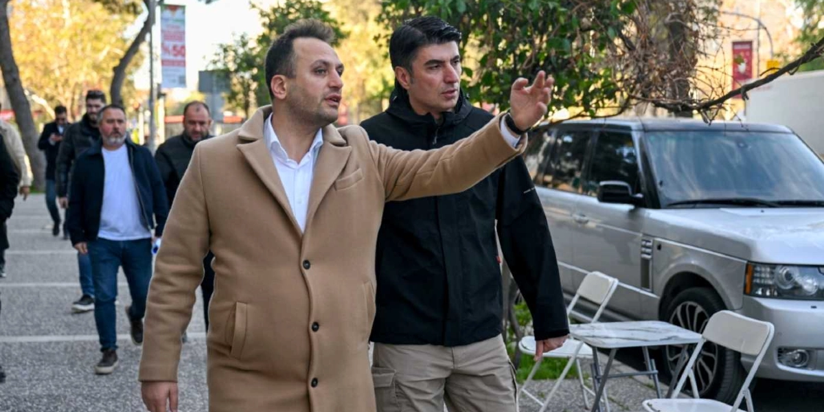 Bej renkli kaban giymiş bir erkek, yanında siyah ceketli bir başka erkekle birlikte dış mekanda yürürken eliyle bir yönü işaret ediyor. Arka planda ise yürüyen diğer erkekler ve park halindeki gümüş renkli bir arazi aracı görülüyor.
