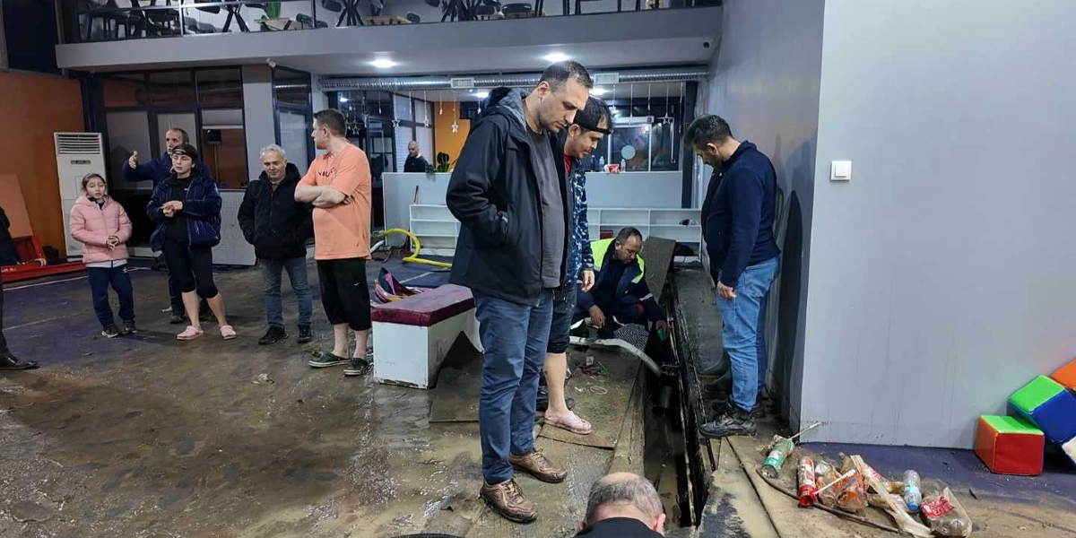 Sel sonrası çamurla kaplanmış bir kapalı alanda, bir grup insan su tahliyesi ve temizlik çalışmalarıyla ilgileniyor. Bir görevli yerdeki bir kanalın içine bakarken, diğerleri çevresinde durarak çalışmaları izliyor. Zemin çamurlu ve ıslak görünürken, köşede çocuk oyun alanına ait renkli küpler ve yerde bazı atıklar bulunuyor.