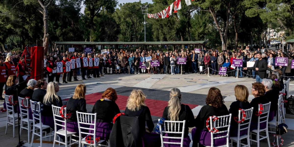 Açık havada, park alanında toplanmış kalabalık bir grup kadın, 8 Mart Dünya Kadınlar Günü kapsamında bir etkinlik gerçekleştiriyor. Ön planda, siyah üst ve mor etek giymiş, ellerinde tef tutan bir grup kadın beyaz sandalyelerde yarım daire şeklinde oturuyor. Arka planda ayakta duran katılımcılar, üzerinde 