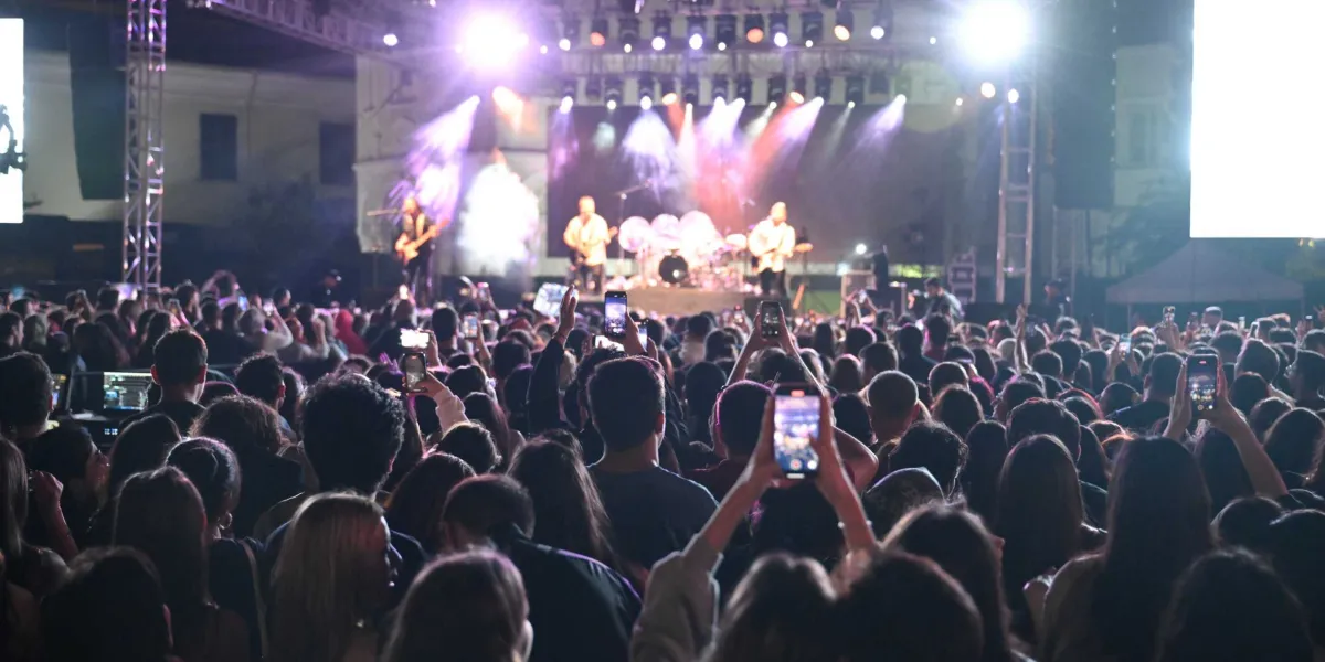 Gece vakti açık havada düzenlenen bir konserde, sahnedeki müzik grubunu izleyen kalabalık bir genç topluluğu görülüyor. Sahne, parlak ışıklar ve duman efektleriyle aydınlatılmış durumda. İzleyicilerin çoğu ellerinde telefonlarıyla sahneyi kaydediyor veya fotoğraf çekiyor; kalabalığın başları ve havaya kaldırılmış telefon ekranları sahneye doğru dönük.