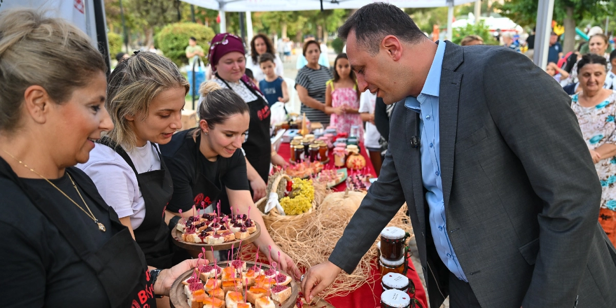Açık havada kurulan bir stantta, siyah önlük giymiş üç kadın, üzerinde küçük kanepeler bulunan ahşap tepsileri uzatarak ikramda bulunuyor. Takım elbiseli bir erkek, standın önünde durarak tepsiden bir kanepe alıyor. Masanın üzerinde çeşitli reçel kavanozları, süslemeler ve doğal ürünler yer alıyor; arka planda ise etkinliği izleyen diğer insanlar görülüyor.