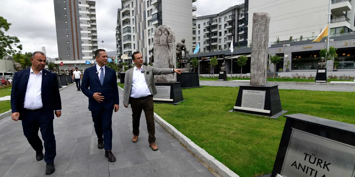 Modern binaların önündeki park alanında yürüyen üç erkekten biri, eliyle Tonyukuk Yazıtı'nı işaret ederek yanındaki kişiye bir şeyler anlatıyor. Parkta, üzerinde bilgilendirme plakaları bulunan çeşitli tarihi anıtlar ve heykeller yer alıyor. Ön planda 