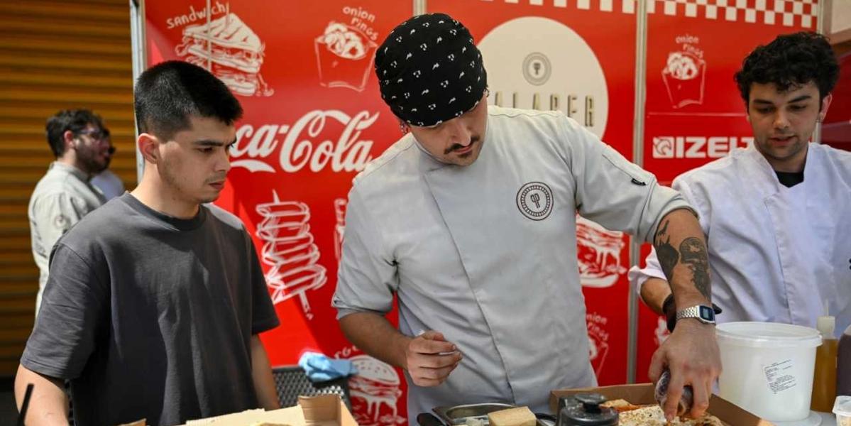 Kırmızı renkli, üzerinde Coca-Cola logosu ve çeşitli yiyecek çizimlerinin bulunduğu bir standın önünde, şef kıyafeti giymiş dövmeli bir genç, önündeki kutuda bulunan yiyeceğe sos ekliyor. Şefin yanında siyah tişörtlü bir genç onu dikkatle izlerken, sağ tarafta beyaz şef önlüğü giymiş başka bir genç daha bulunuyor.