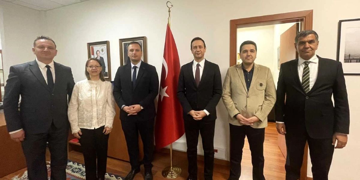 Resmi bir ofis ortamında, arkalarında asılı Türk bayrağı ve duvardaki çerçeveli fotoğrafların önünde yan yana duran, aralarında Bornova Belediye Başkanı Ömer Eşmi'nin de bulunduğu altı kişilik heyet. Gruptaki dört erkek takım elbiseli, bir erkek bej renkli ceketli, bir kadın ise beyaz bluz ve siyah pantolon giymiş şekilde kameraya gülümseyerek poz veriyor.