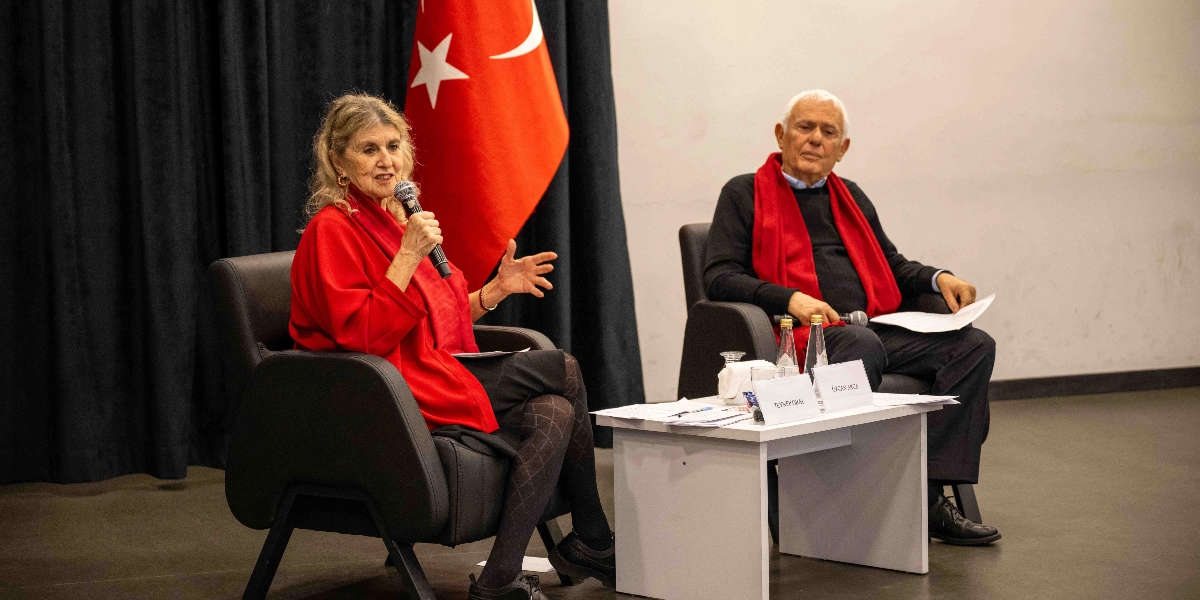 Sahnede yan yana siyah koltuklarda oturan Zeynep Oral ve Özcan Arca, Nazım Hikmet'i anma etkinliğinde konuşma yapıyor. Zeynep Oral kırmızı hırkasıyla elinde mikrofon tutarak konuşurken, yanında oturan Özcan Arca kırmızı atkısıyla onu dinliyor. Önlerinde üzerinde isimliklerin bulunduğu beyaz bir masa, arkalarında ise siyah perde önünde asılı Türk bayrağı yer alıyor.