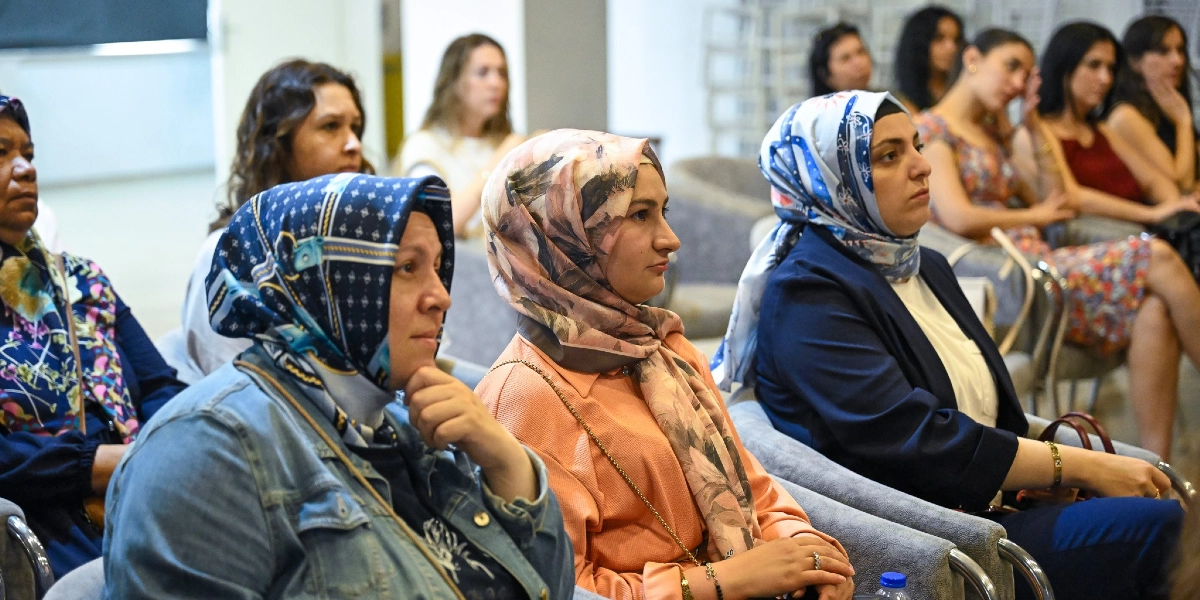 Kapalı bir salonda düzenlenen çocuk eğitimi konulu semineri dikkatle dinleyen, çoğunluğu başörtülü kadınlardan oluşan bir izleyici grubu. Ön planda yan yana oturan üç kadın, arkalarında ise diğer katılımcılar yer alıyor.