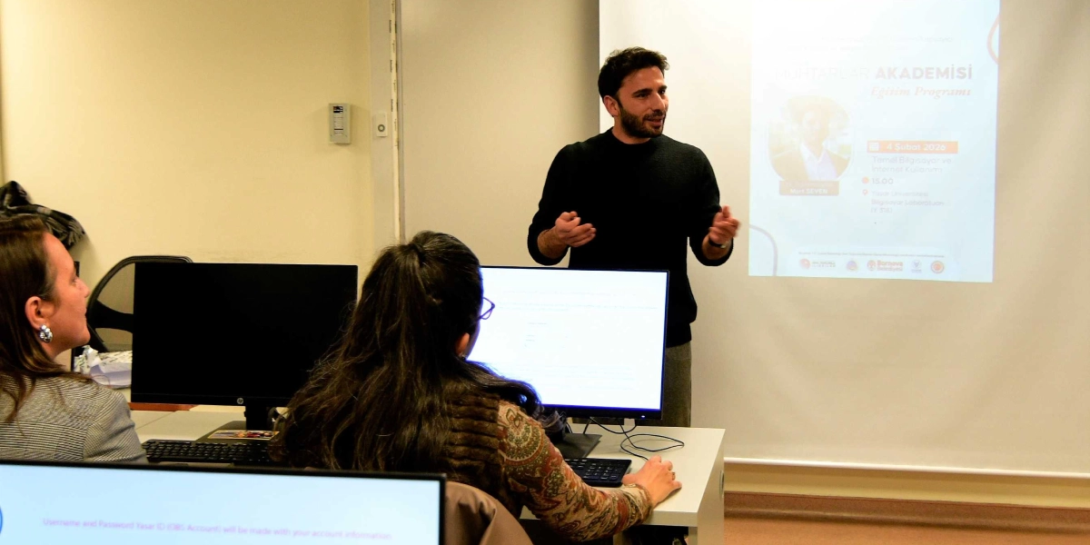 Siyah kazaklı bir eğitmen, bilgisayar laboratuvarında katılımcılara sunum yapıyor. Eğitmenin arkasındaki projeksiyon perdesinde 