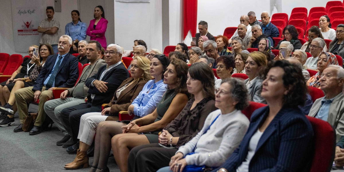 Konferans salonunda kırmızı koltuklarda oturan, farklı yaş gruplarından oluşan kalabalık bir izleyici grubu, sahneye doğru dönmüş dikkatle dinliyor. Katılımcıların çoğu günlük kıyafetler içinde, bazıları not alıyor veya telefonlarıyla ilgileniyor. Salonun sol tarafındaki duvarda Bornova Belediyesi'ne ait 