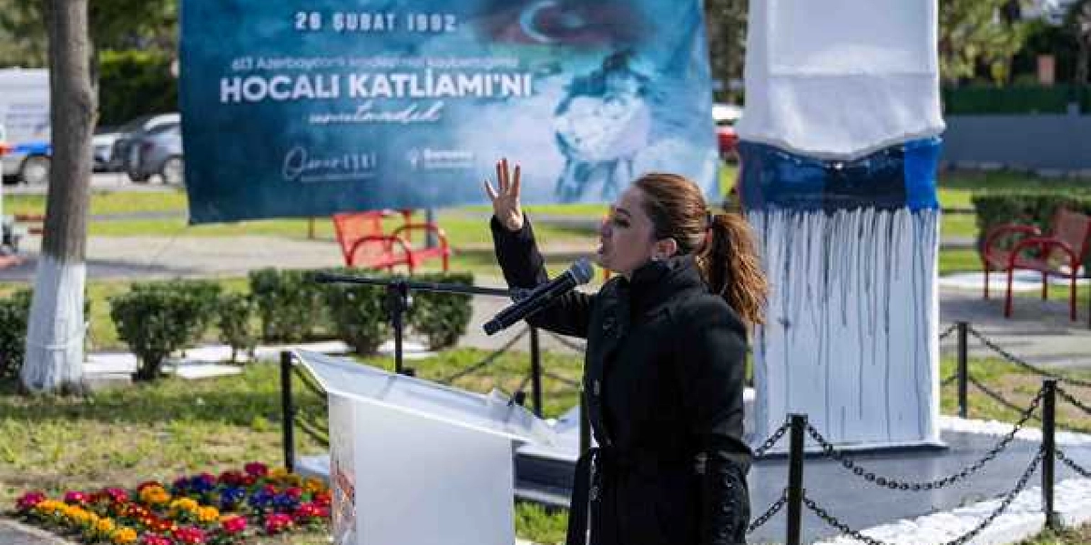 Açık havada düzenlenen bir anma töreninde, kürsüde konuşma yapan bir kadın yer alıyor. Kadın, siyah kaban giymiş ve elini havaya kaldırarak konuşuyor. Arka planda üzerinde 
