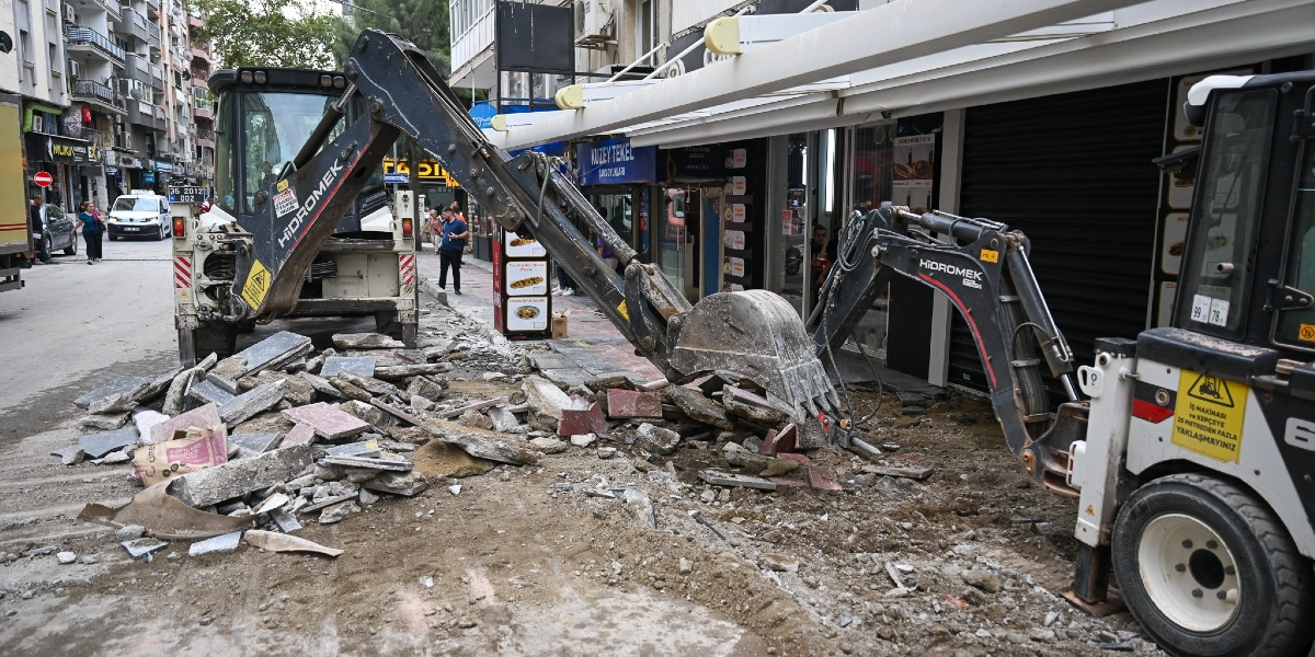 Bornova Zafer Caddesi'nde bir iş makinesi, dükkanların önündeki eski kaldırım taşlarını sökerek çevre düzenleme çalışması yapıyor. İş makinesinin kazıcı kolu, sökülen beton ve taş parçalarının bulunduğu çalışma alanının üzerinde duruyor. Arka planda cadde üzerindeki dükkanlar, yayalar ve trafik akışı görülüyor.