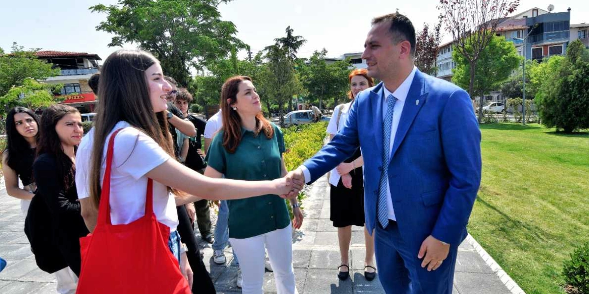 Mavi takım elbiseli bir erkek, açık havada bir parkta genç bir kadınla tokalaşıyor. Genç kadının omzunda kırmızı bir bez çanta bulunuyor. İkilinin yanında, onları izleyen başka gençler ve yeşil gömlekli bir kadın yer alıyor. Arka planda ağaçlar, çimenlik bir alan ve binalar görülüyor.