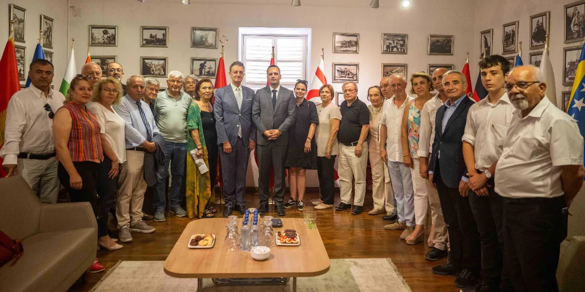 Kapalı bir mekanda, duvarları tarihi fotoğraflarla süslü bir odada bir araya gelmiş, farklı yaş gruplarından oluşan kalabalık bir grup insan. Grubun merkezinde, takım elbiseli iki erkek yan yana duruyor; etraflarında ise resmi ve günlük kıyafetli kadın ve erkekler yer alıyor. Arka planda çeşitli ülke bayrakları ve ortada bir sehpa üzerinde ikramlıklar bulunuyor. Genel atmosfer resmi ve toplu bir buluşmayı yansıtıyor.