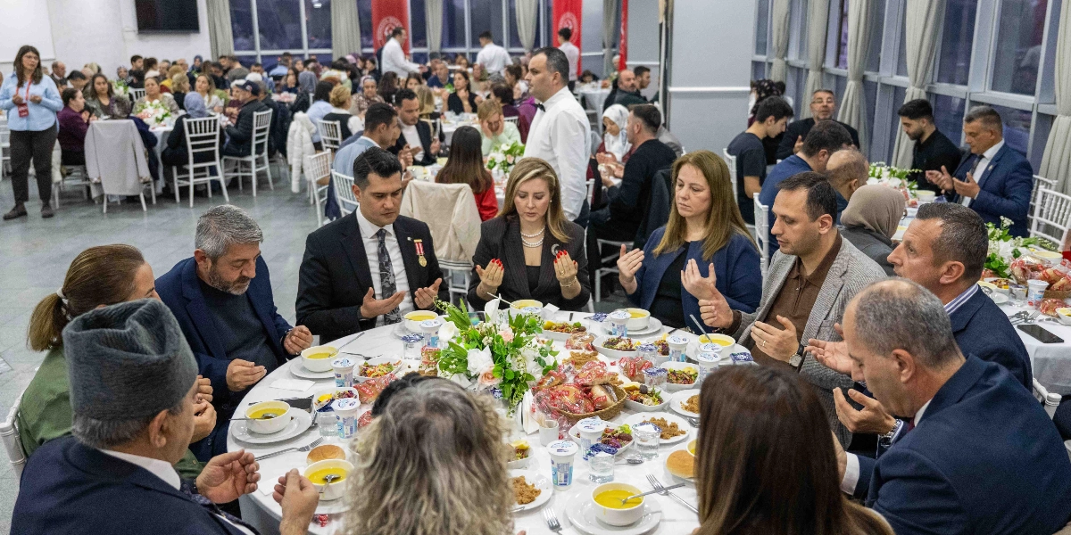 Geniş bir salonda beyaz örtülü yuvarlak masaların etrafında toplanmış kalabalık bir grup insan, önlerindeki yemeklerden önce ellerini açarak dua ediyor. Ön plandaki masada oturan kadın ve erkekler, masanın ortasındaki çiçek aranjmanı ve çeşitli yemek tabakları eşliğinde dua ederken görülüyor. Arka planda ise benzer şekilde yemek masalarında oturan çok sayıda davetli yer alıyor.
