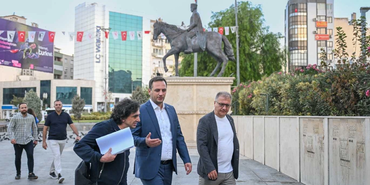 Bornova Cumhuriyet Meydanı'nda, arka planda Atatürk'ün at üzerindeki heykeli ve çevredeki binaların önünde yürüyen üç erkek. Öndeki grupta yer alan lacivert ceketli bir kişi yanındaki arkadaşına bir şeyler anlatırken, yanındaki gri ceketli kişi onu dinliyor. Grubun hemen önünde elinde dosyalar taşıyan bir başka kişi daha yürüyor. Arka planda Türk bayrakları ve şehir dokusu görülüyor.