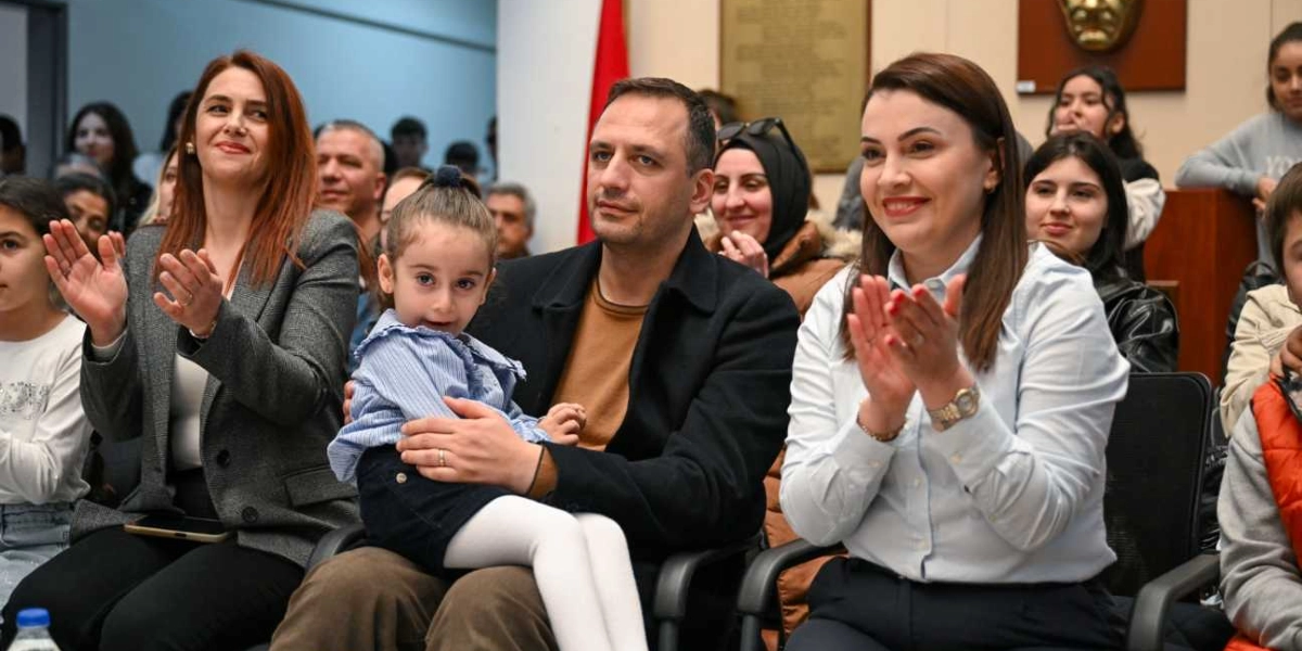 Kapalı bir salonda düzenlenen etkinlikte, izleyiciler arasında oturan bir grup insan alkışlıyor. Ön planda, kucağında küçük bir kız çocuğu tutan bir adam ve yanında oturan, gülümseyerek alkışlayan iki kadın yer alıyor. Arka planda ise etkinliği takip eden diğer katılımcılar bulunuyor.