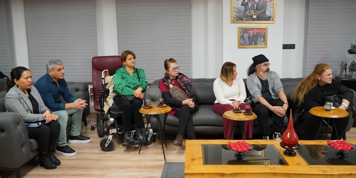Modern bir ofis ortamında gri koltuklarda yan yana oturmuş, dikkatle dinleyen yedi kişilik bir grup. En solda gri ceketli bir kadın ve yanında mavi gömlekli bir erkek, hemen yanlarında tekerlekli sandalyede oturan yeşil gömlekli bir kadın bulunuyor. Grubun devamında bordo bluzlu, beyaz üstlü, siyah şapkalı ve uzun saçlı bir erkek ile siyah kıyafetli bir kadın yer alıyor. Önlerinde ahşap bir orta sehpa ve üzerinde küçük ikram tabakları ile çay bardakları duruyor. Arka duvarda ise çerçeveli fotoğraflar asılı.