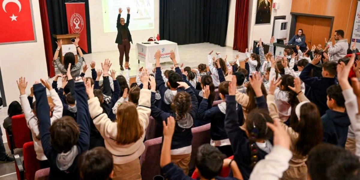 Bir konferans salonunda, sahnedeki eğitmenle birlikte kollarını havaya kaldırmış ilkokul ve ortaokul öğrencileri. Sahnenin solunda Türk bayrağı ve Bornova Belediyesi flaması yer alırken, eğitmen arkasındaki yansıtıcı ekranda sunum eşliğinde öğrencilere egzersiz yaptırıyor. Öğrenciler koltuklarında oturarak veya ayakta, eğitmeni takip ederek aktif bir şekilde eğitime katılıyor.