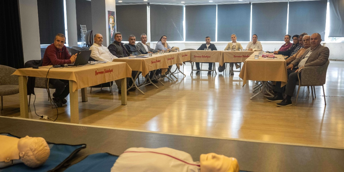 Geniş bir salonda, U düzeninde birleştirilmiş masaların etrafında oturan bir grup muhtar ve katılımcı, önlerinde duran ilk yardım mankenlerine bakarak eğitim alıyor. Masaların üzerinde Bornova Belediyesi yazılı örtüler serili ve katılımcılar dikkatle dinliyor. Ön planda, eğitimde kullanılan iki adet ilk yardım mankeni net bir şekilde görülüyor.