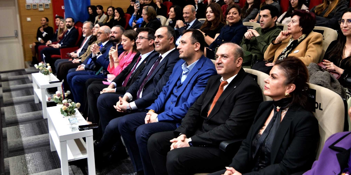 Konferans salonunda yan yana oturmuş, resmi kıyafetli bir grup insan izleyici koltuklarında bir etkinliği takip ediyor. Ön sırada, koyu renkli takım elbiseli erkekler ve şık giyimli kadınlar dikkatle karşıya bakıyor; aralarında Bornova Belediye Başkanı Mustafa İduğ'un da bulunduğu grup, eğitimle ilgili bir toplantıda yer alıyor. Salonun genelinde kalabalık bir katılımcı kitlesi bulunuyor ve önlerindeki beyaz masaların üzerinde küçük saksı çiçekleri yer alıyor.
