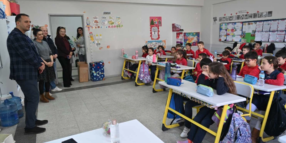 Sınıf ortamında, kırmızı üniformalı ilkokul öğrencileri sıralarında otururken, sol tarafta ayakta duran bir grup yetişkin onlara hitap ediyor. Öğrenciler dikkatle ön tarafa bakarken, sınıfın duvarları çocukların yaptığı resimler ve eğitim materyalleriyle süslenmiş durumda.