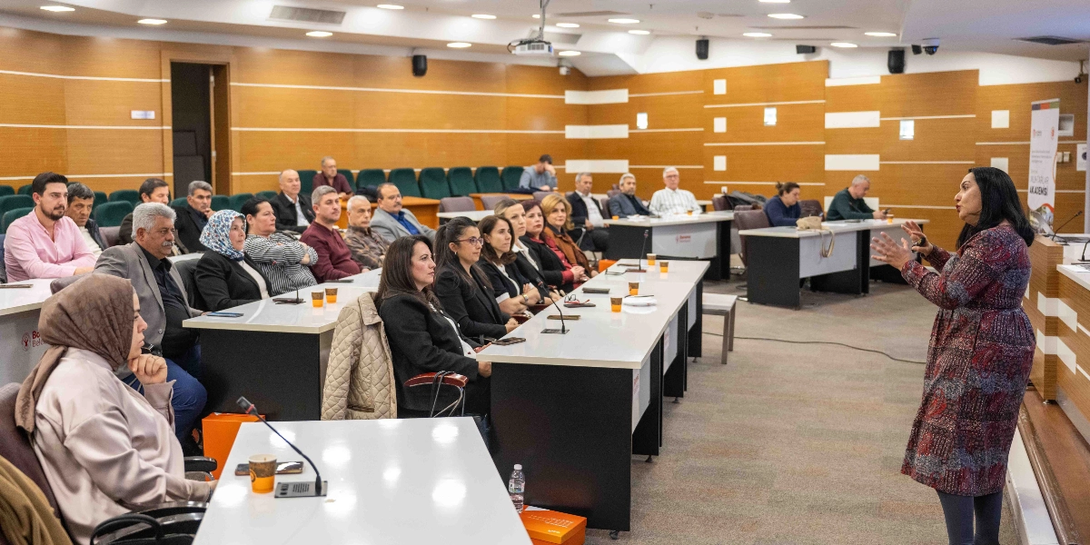Modern bir konferans salonunda, Bornova Muhtarlar Akademisi kapsamında düzenlenen bir eğitimde, ayakta duran bir konuşmacı katılımcılara hitap ediyor. U-şeklindeki masalarda oturan çok sayıda muhtar ve katılımcı, konuşmacıyı dikkatle dinliyor. Salonun duvarları ahşap panellerle kaplı ve masaların üzerinde mikrofonlar ile içecek bardakları bulunuyor. Sağ tarafta üzerinde 