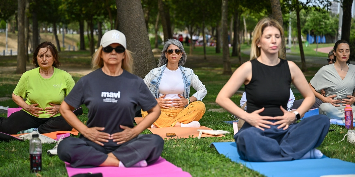 Ağaçlık bir parkta, çimenlerin üzerine serilmiş matların üzerinde bağdaş kurarak oturan ve ellerini karınlarının üzerine koymuş nefes egzersizi yapan beş kadın. Kadınlar rahat kıyafetler içinde, gözleri kapalı ve odaklanmış bir şekilde meditasyon yapıyorlar.