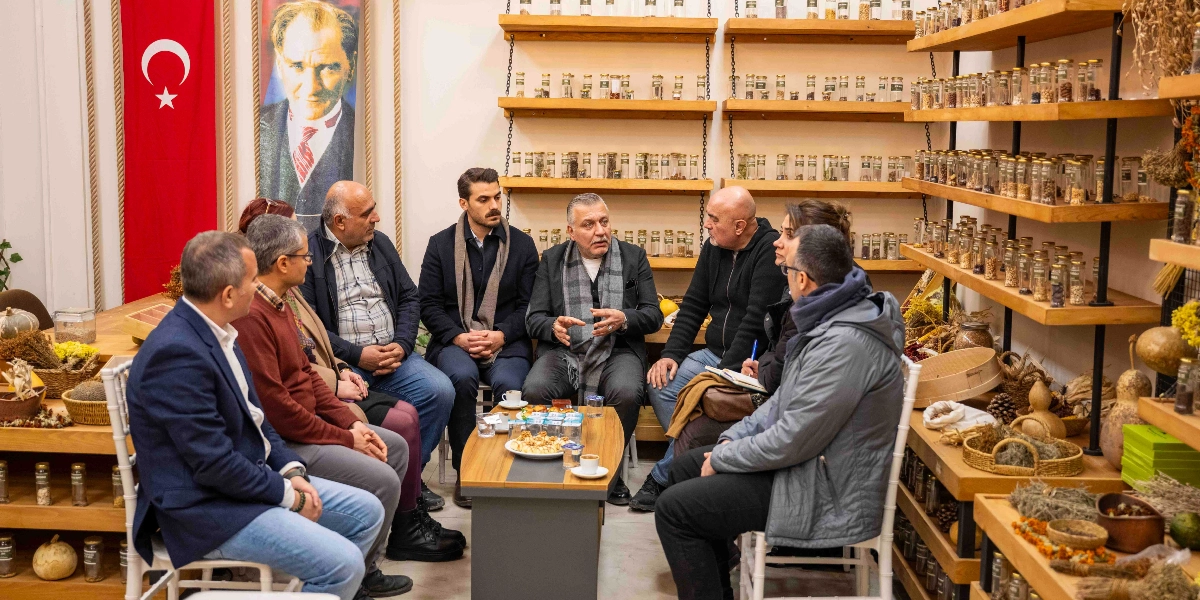 Bornova'da onarıcı tarım üzerine yapılan bir toplantıda, bir masa etrafında toplanmış sekiz kişilik bir grup yer alıyor. Arka planda duvarda asılı Türk bayrağı ve Mustafa Kemal Atatürk portresi bulunuyor. Duvar boyunca uzanan ahşap raflarda, içlerinde çeşitli tohum ve bitkilerin olduğu çok sayıda cam kavanoz dizili. Grup üyeleri, aralarında konuşarak toplantıya devam ediyor; masanın üzerinde ise küçük ikramlıklar ve kahve fincanları yer alıyor.