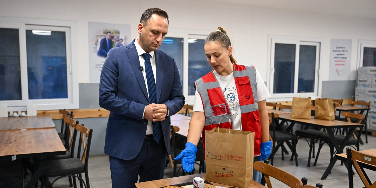 Lacivert takım elbiseli bir erkek ve üzerinde Bornova Belediyesi logolu kırmızı bir yelek bulunan, mavi eldivenli bir kadın görevli, masanın üzerindeki beslenme paketlerini inceliyor. Kadın görevli, üzerinde Bornova Belediyesi ve Öncü Kent Bornova yazılı kağıt bir çantayı işaret ediyor; masanın üzerinde ise öğrencilere dağıtılmak üzere hazırlanan yiyecek ve içecek paketleri bulunuyor. Arka planda ise yemekhane düzeninde masalar ve sandalyeler yer alıyor.