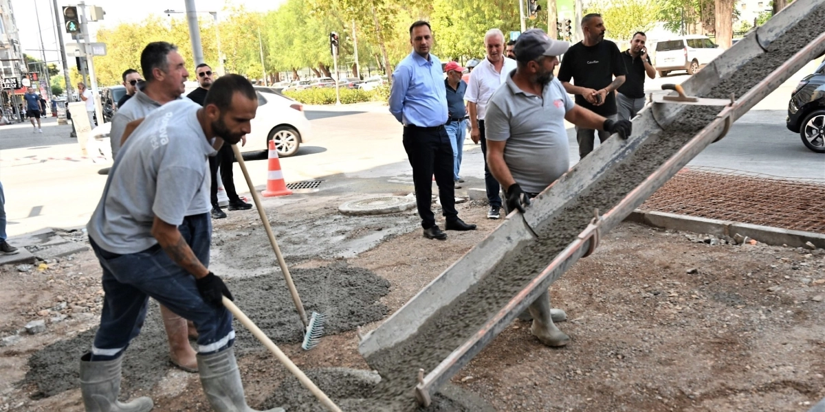 Bornova Belediyesi logolu gri tişört giyen işçiler, bir beton mikserinden dökülen harcı kürek ve tırmık yardımıyla yol kenarındaki alana yayıyor. Çalışma alanının çevresinde, aralarında resmi kıyafetli yetkililerin de bulunduğu bir grup insan çalışmaları izliyor. Arka planda ağaçlıklı bir cadde, park halindeki araçlar ve trafik ışıkları görülüyor.