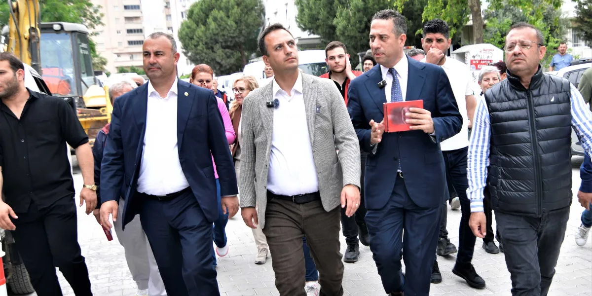 Açık havada, parke taşlı bir yolda yan yana yürüyen bir grup erkek; ön planda, koyu renk takım elbiseli, gri ceketli ve siyah yelekli kişilerin ağırlıkta olduğu bir heyet yer alıyor. Ortadaki lacivert takım elbiseli kişi elinde kırmızı bir kitap tutuyor ve konuşuyor. Arka planda ise yürüyüşe katılan diğer insanlar ve inşaat çalışmaları için kullanılan bir iş makinesi görünüyor.