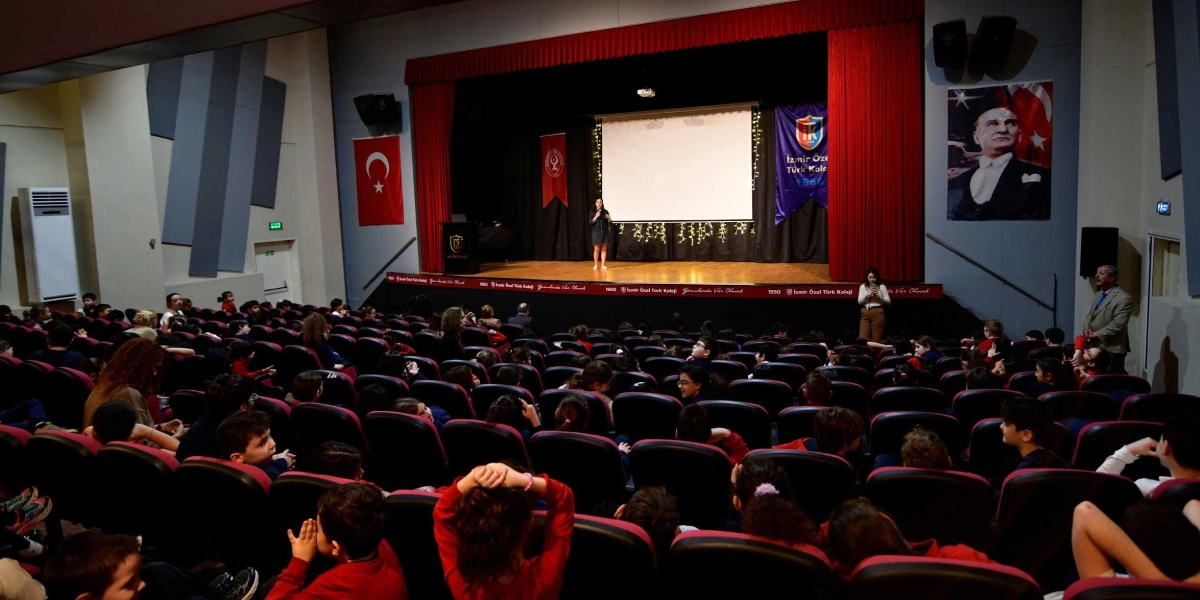 Bir okulun konferans salonunda, sahne üzerinde konuşma yapan bir kadın ve onu dinleyen çok sayıda ilkokul ve ortaokul öğrencisi bulunuyor. Sahnenin arka planında beyaz bir projeksiyon perdesi, yanında Türk bayrağı, İzmir Özel Türk Koleji flaması ve Mustafa Kemal Atatürk'ün portresi yer alıyor. Sahne önünde Bornova Belediyesi tarafından düzenlenen sağlıklı yaşam eğitimi kapsamında öğrenciler ilgiyle sunumu takip ediyor.