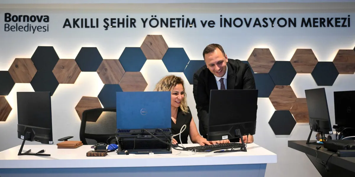 Bornova Belediyesi Akıllı Şehir Yönetim ve İnovasyon Merkezi'nde, beyaz bir çalışma masasının başında oturan sarı saçlı bir kadın ve onun yanında ayakta durarak bilgisayar ekranını inceleyen takım elbiseli bir erkek, birlikte çalışırken gülümseyerek ekrana bakıyorlar. Arka planda, üzerinde Bornova Belediyesi logosu ve 