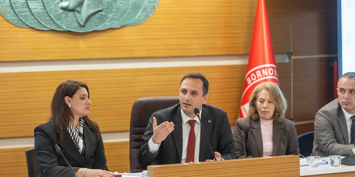 Bornova Belediyesi meclis salonunda, ahşap kaplamalı duvarın önünde, üzerinde Bornova Belediyesi logosu bulunan kırmızı bir bayrağın yanında dört kişi oturuyor. Ortada konuşma yapan, siyah takım elbiseli ve kırmızı kravatlı bir erkek, yanında ise siyah ceketli bir kadın, kahverengi ceketli başka bir kadın ve gri ceketli bir erkek yer alıyor. Masaların üzerinde mikrofonlar bulunuyor.