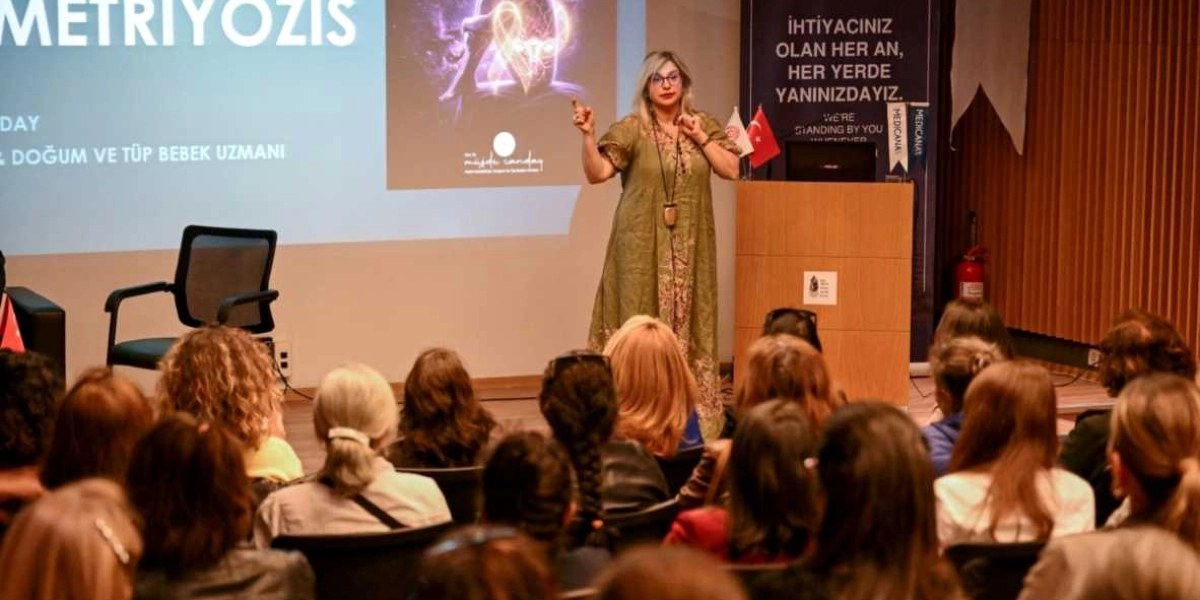 Bir konferans salonunda, kürsü önünde duran sarı saçlı ve gözlüklü bir kadın konuşmacı, izleyicilere endometriozis hakkında sunum yapıyor. Konuşmacı, üzerinde desenli, uzun, haki renkli bir elbise ile ellerini kullanarak açıklamalarda bulunuyor. Arkasındaki dev ekranda 