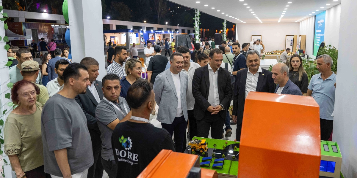 İzmir Enternasyonal Fuarı'nda Bornova Belediyesi standında bir grup insan, önlerinde yer alan şehir planlama maketini incelemektedir. Maketin başında Bornova Belediye Başkanı Mustafa İduğ ve beraberindeki heyet yer alırken, çevredeki ziyaretçiler de projeye ilgi göstermektedir. Standın arka planında ise fuar alanındaki diğer stantlar ve kalabalık bir ziyaretçi grubu görülmektedir.