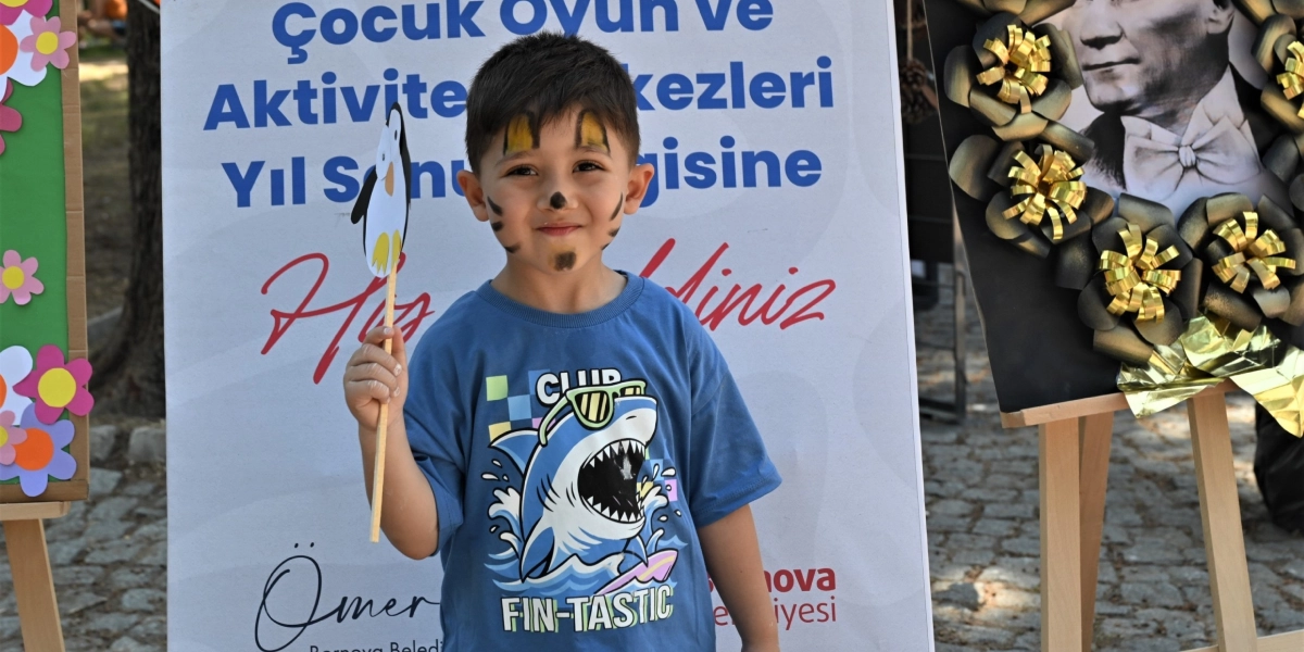 Yüzü kaplan şeklinde boyanmış, üzerinde köpek balığı baskılı mavi bir tişört olan küçük bir erkek çocuk, elinde tuttuğu penguen figürlü bir çubukla gülümsüyor. Çocuğun arkasında 