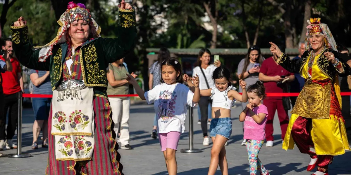 Açık havada düzenlenen bir etkinlikte, geleneksel kıyafetler giymiş iki kadın ve onlara eşlik eden üç küçük kız çocuğu halay çekiyor. Sol taraftaki kadın koyu yeşil, altın işlemeli bir kıyafet ve başlık takarken, sağ taraftaki kadın sarı ve siyah renkli benzer bir geleneksel kostüm giyiyor. Çocuklar ise günlük kıyafetleriyle neşeyle dansa katılıyor. Arka planda etkinliği izleyen insanlar ve ağaçlıklı bir park alanı görülüyor.