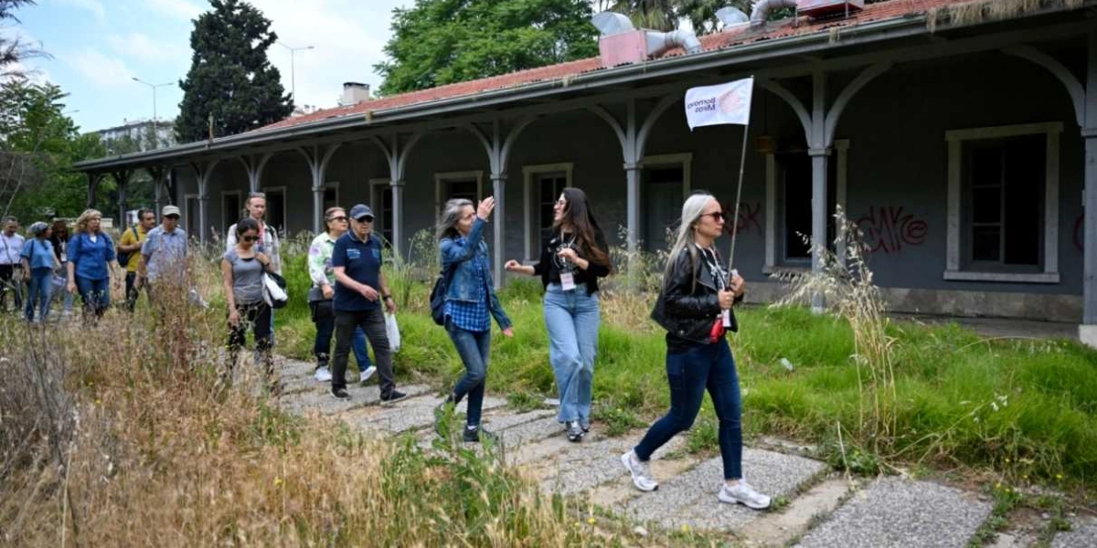 Tarihi bir tren istasyonu binasının önündeki taşlı yolda yürüyen bir grup insan. En önde, üzerinde Bornova Miras yazılı beyaz bir bayrak taşıyan kadın rehberlik ediyor. Grubun geri kalanı, otlarla kaplı patikada ilerleyerek tarihi yapıyı inceliyor.