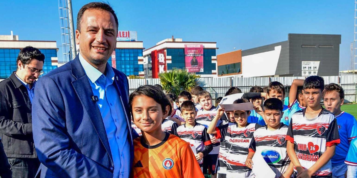 Mavi ceketli bir yetişkin, futbol forması giymiş çocuklardan oluşan bir grupla açık havada yan yana duruyor. Ön planda, turuncu bir kaleci forması giyen genç bir çocuk gülümsüyor. Arkasında ise beyaz, siyah ve kırmızı renkli futbol formaları giymiş, ellerinde belgeler tutan diğer çocuklar yer alıyor. Arka planda modern binalar ve açık mavi bir gökyüzü görülüyor.