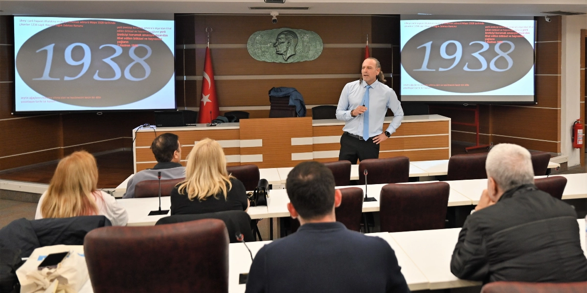 Modern bir konferans salonunda, kürsüde konuşma yapan uzun saçlı bir erkek ve onu dinleyen bir grup katılımcı görülüyor. Konuşmacının arkasındaki iki büyük ekranda, 1938 yılına ait tarım ve hayvancılıkla ilgili yasal düzenlemeleri anlatan metinler ve büyük puntolarla yazılmış 