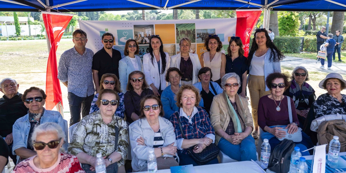 Açık havada, Bornova Belediyesi'nin kurduğu bir çadırın altında, üniversite eğitimi alan yaşlı vatandaşlar ve eğitmenlerin bir arada olduğu toplu fotoğraf. Arka planda üzerinde çeşitli etkinlik fotoğraflarının bulunduğu bir branda yer alıyor. Ön sırada oturan, güneş gözlüklü ve şık giyimli yaşlı kadınlar ile arka sırada ayakta duran belediye yetkilileri ve eğitmenler gülümsüyor. Masaların üzerinde su şişeleri ve Bornova Belediyesi logolu materyaller bulunuyor.