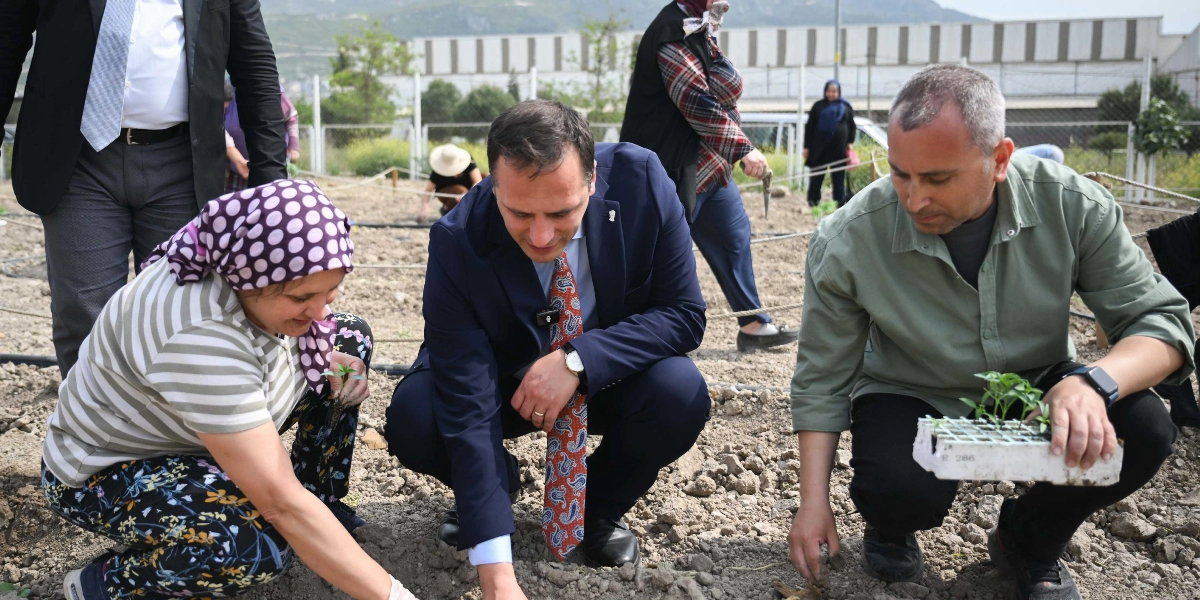 Koyu renk takım elbiseli ve desenli kravatlı bir erkek, yanında çizgili bluzlu ve başörtülü bir kadın ile haki renkli gömlekli bir erkekle birlikte toprak bir alanda diz çökmüş, fide dikimi yapıyor. Haki gömlekli adam elinde içinde küçük fideler bulunan bir viyol tutuyor. Arka planda ise tarım alanında çalışan diğer insanlar ve bir tel çit görülüyor.