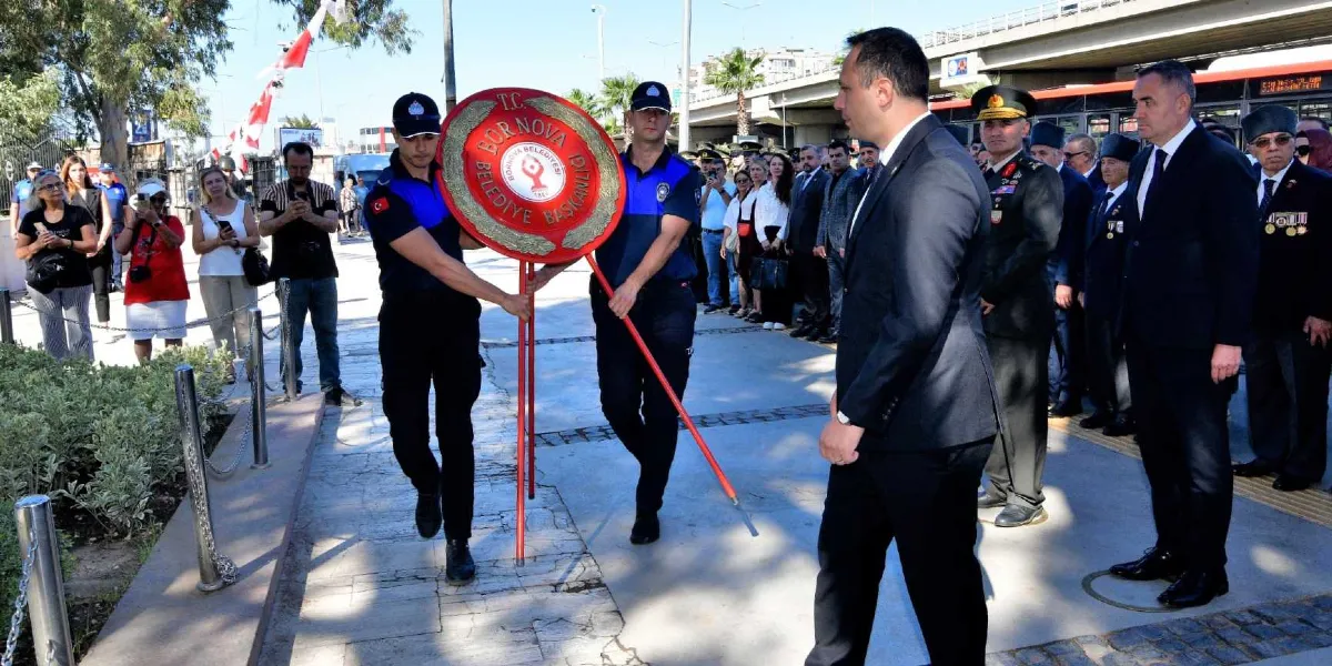 Bornova Belediyesi'ne ait kırmızı renkli bir çelengi taşıyan iki polis memuru, tören alanındaki anıta doğru ilerliyor. Onların önünde ve arkasında, aralarında askeri yetkililerin ve sivil protokol üyelerinin bulunduğu bir grup insan, resmi bir tören ciddiyetiyle duruyor. Arka planda ise cep telefonlarıyla çekim yapan vatandaşlar ve ağaçlar görülüyor.
